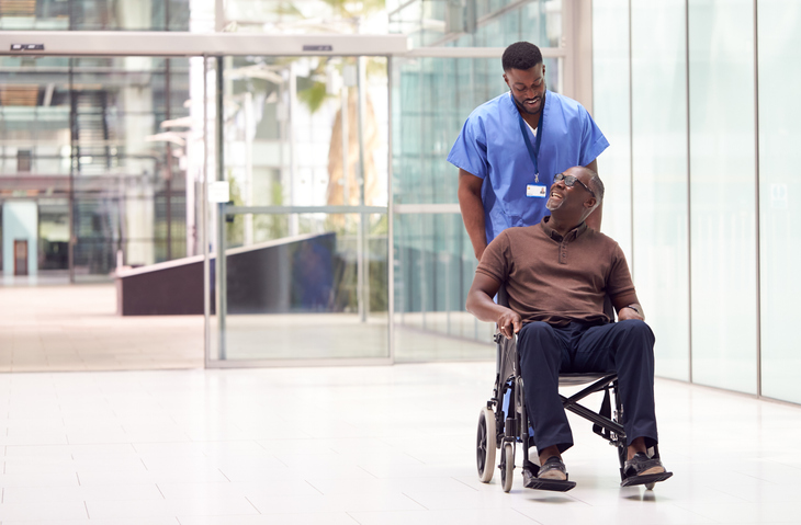 A healthcare worker in blue scrubs pushes a smiling man in a wheelchair through a bright, modern building with glass walls, highlighting the positive impact of a personal wheelchair budget on his independence and comfort.