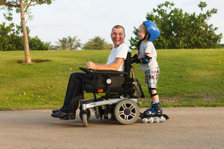 A smiling man in a wheelchair, enjoying the freedom his personal wheelchair budget provides, is pushed by a young boy on rollerblades wearing a blue helmet, outdoors on a sunny day with green grass and trees in the background.