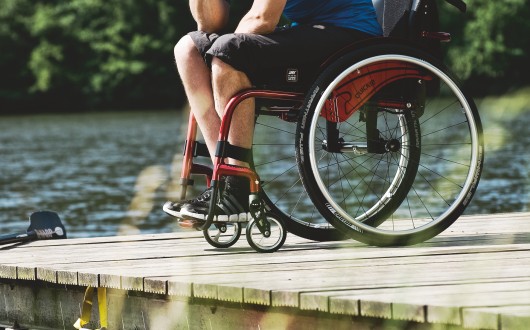 A person in a Quickie Argon 2 wheelchair sits on a wooden jetty by a lake, with trees in the background. Only their legs and wheelchair are visible.