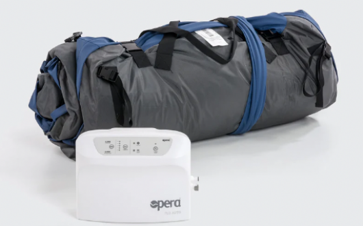 A rolled-up grey and blue sleeping mat sits next to a white electronic device labelled Opera Flo Auto Air. The device features buttons and indicators on top. Both items are placed on a light surface.