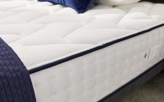 Close-up of the Opera Gel 1000 Mattress, showcasing its white quilted pattern and navy blue piping along the edge. Part of a navy blue blanket and blue pillows are visible at the top.