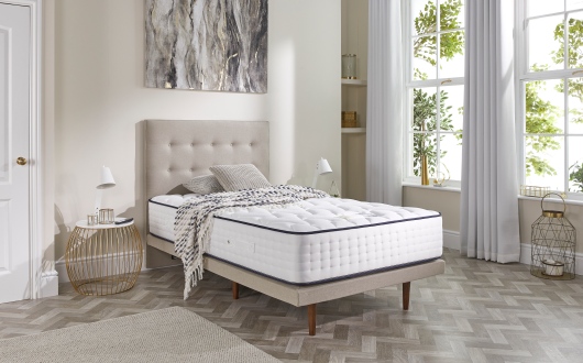 A modern bedroom featuring a beige upholstered bed with the luxurious Opera Gel Pro 4000 mattress, complemented by plush pillows, a cosy throw, round bedside tables with elegant lamps, large windows draped in white curtains, and abstract wall art. Sunlight brightens the room.