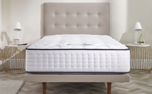 A neatly made bed with a thick, quilted white Opera Gel Pro 4000 mattress sits on a beige upholstered frame. Two round bedside tables with lamps are on either side, against a neutral wall.
