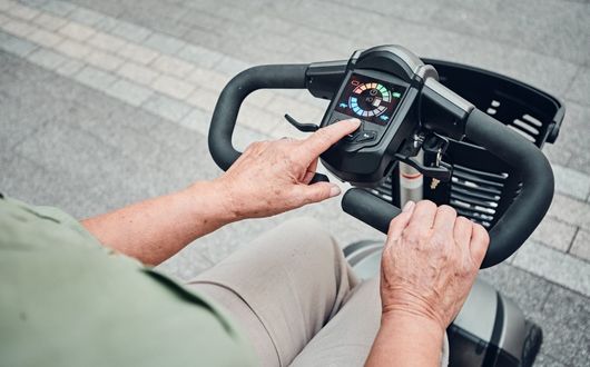 A person operates a Motion Healthcare Aura mobility scooter, pressing a button on the digital control panel. The scooter’s handlebars and basket are visible, with a paved path in the background.