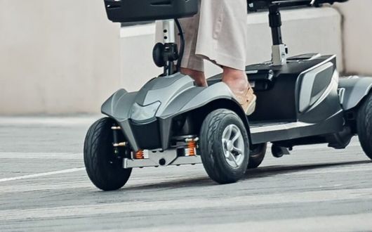 Close-up of a person’s legs and feet on the footrest of a Motion Healthcare Aura mobility scooter, with the scooter’s front wheels and part of its frame visible on a paved surface.