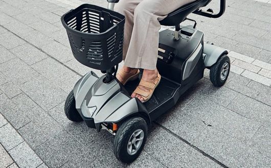 A person wearing beige sandals and light trousers sits on a Motion Healthcare Aura mobility scooter with a black basket, parked on a paved pavement.
