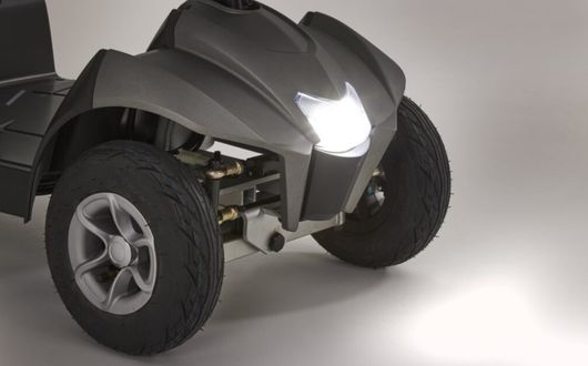 Close-up of the front wheels and illuminated headlamp of the Motion Healthcare Aura, a small, black, four-wheeled vehicle, against a light background.