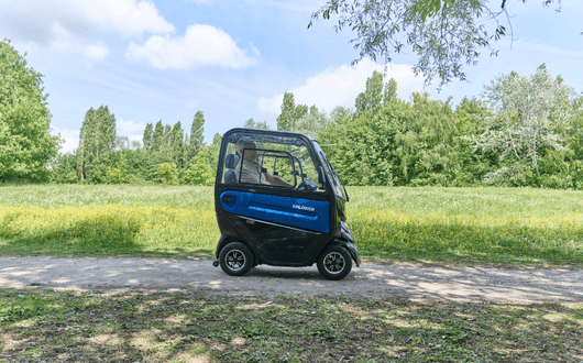 A blue Xplorer mobility scooter with a protective cab is parked on a path in a sunny, green park with trees and grass in the background.