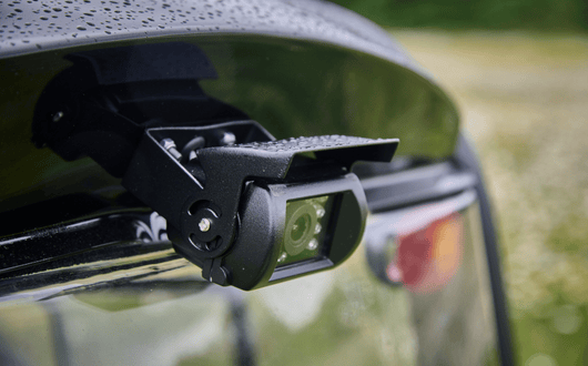 Close-up of an Xplorer backup camera mounted on the rear of a vehicle, with water droplets visible on the surface, likely after rain. The background is blurred with greenery and part of a red tail light visible.