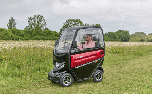 An older adult sits inside a small, enclosed Xplorer mobility scooter with black and red panels, driving through a grassy field with trees in the background.