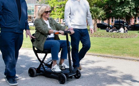 A smiling woman rides a small second-hand mobility scooter in a park alongside two men walking. There is grass, trees, and people sitting on the lawn in the background.