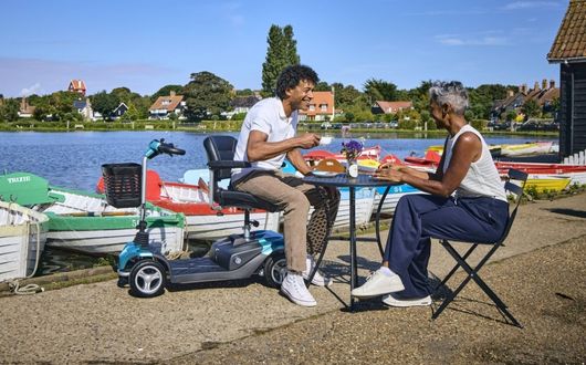 Two older adults sit at a small table by a lakeside, talking and smiling. One is seated on a used mobility scooter, enjoying the scenic view. Colourful boats and houses are visible in the background under a blue sky.