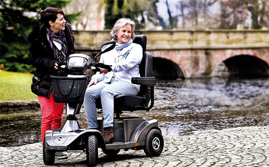 Two women outdoors by a river: one stands smiling whilst the other, older woman sits on a second-hand mobility scooter. There is a stone bridge and trees in the background.