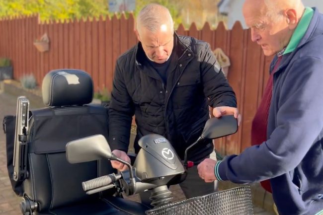 Two older men stand outdoors next to a used mobility scooter. One man is adjusting the scooter’s wing mirror while the other watches closely. There is a wooden fence and greenery in the background.