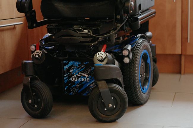 Close-up of the wheels and base of a black and blue Quickie powered wheelchair, similar to a used mobility scooter, on a tiled kitchen floor. The wheelchair is positioned near wooden cupboards.