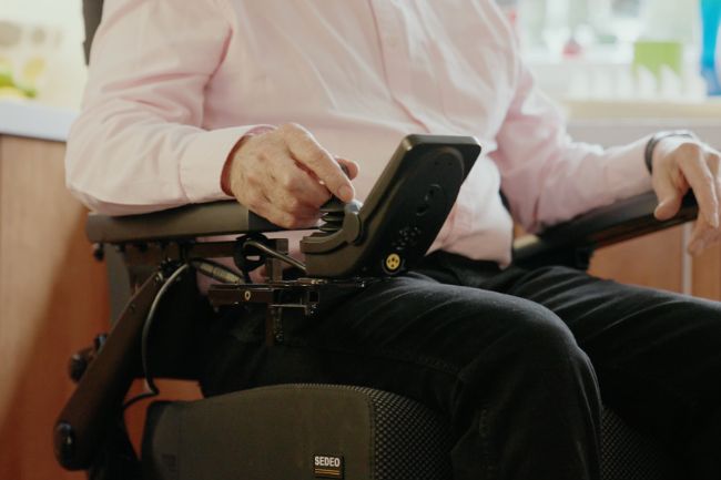 A person in a pink shirt uses a joystick to operate a used mobility scooter converted into a powered wheelchair indoors.